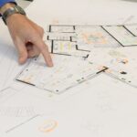 Eine Hand zeigt auf einen detaillierten Bauplan auf einem Tisch mit verschiedenen Zeichnungen.