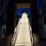 Ein beleuchteter Treppenaufgang führt durch einen rechteckigen Rahmen in die Dunkelheit.