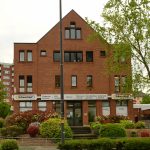 Rotes Backsteingebäude mit Schildern für Elektrotechnik, umgeben von bunten Pflanzen und Bäumen.